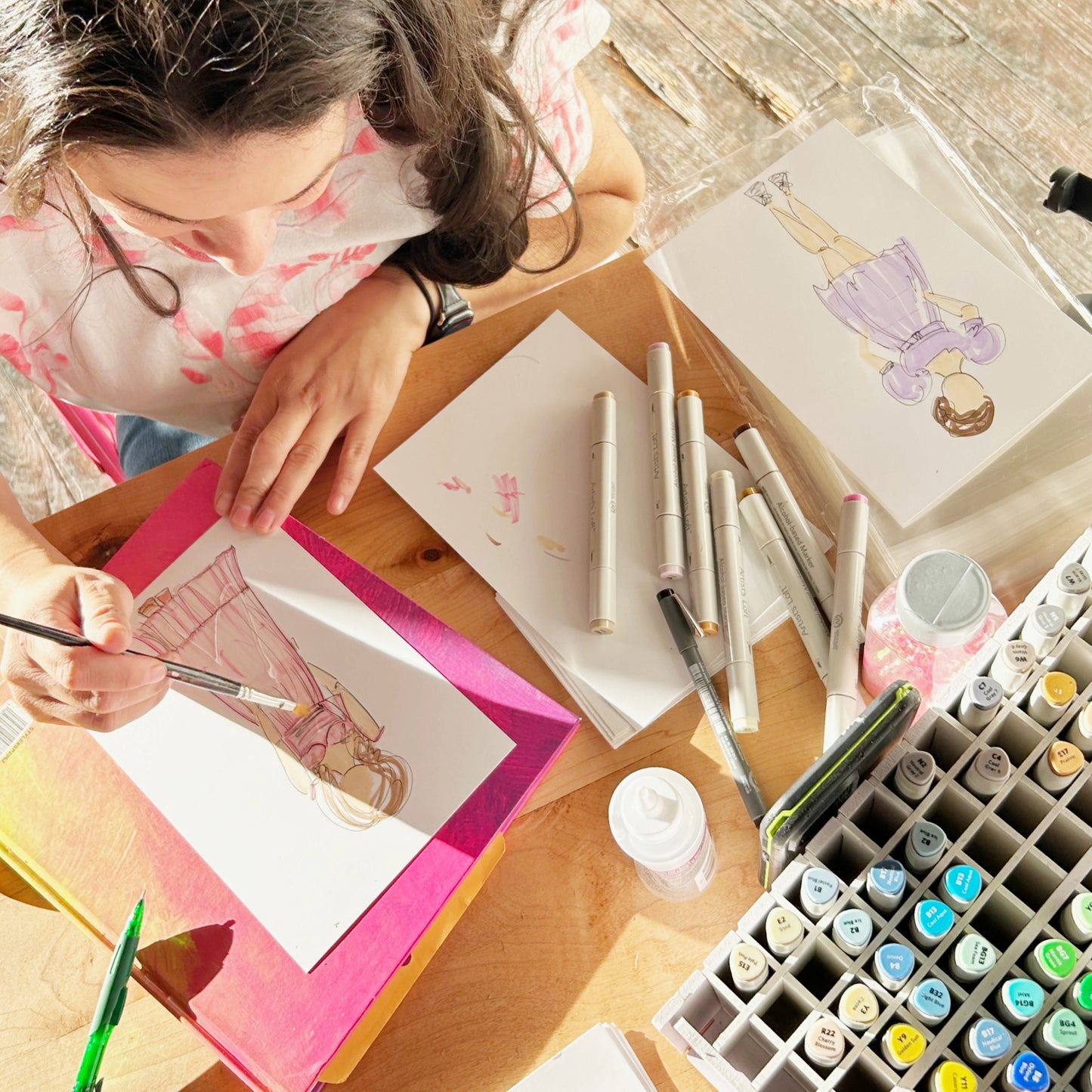 Artist Natalie Zoghby working on a custom guest portrait at a live event, surrounded by markers and pens on a wooden table. The drawing captures a guest’s outfit and essence with intricate pen and ink details.