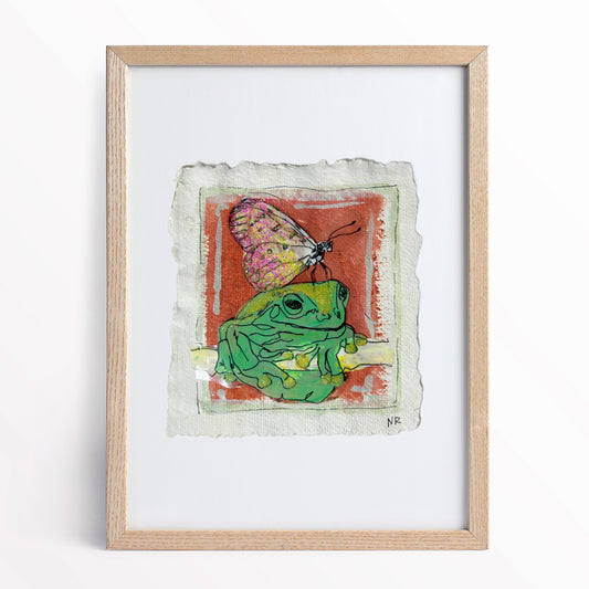 5.5x6 inch painting of a frog with a butterfly by Natalie Russo, framed and displayed on a minimalist counter. The acrylic artwork on handmade watercolor paper adds a whimsical touch to the modern space.