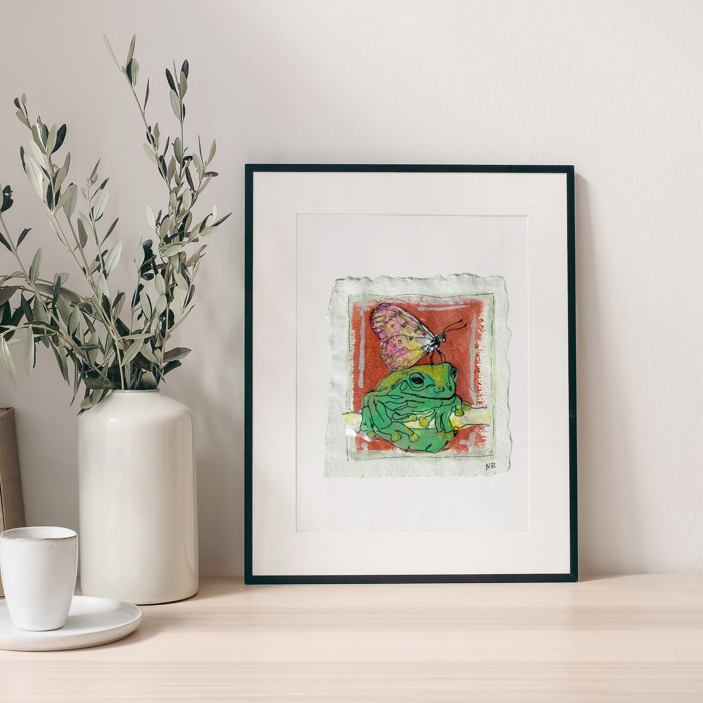 Framed 5.5x6 inch Frog and Butterfly painting by Natalie Russo, featuring a green frog and a butterfly against a warm red background. The handmade watercolor paper adds texture to the acrylic artwork.