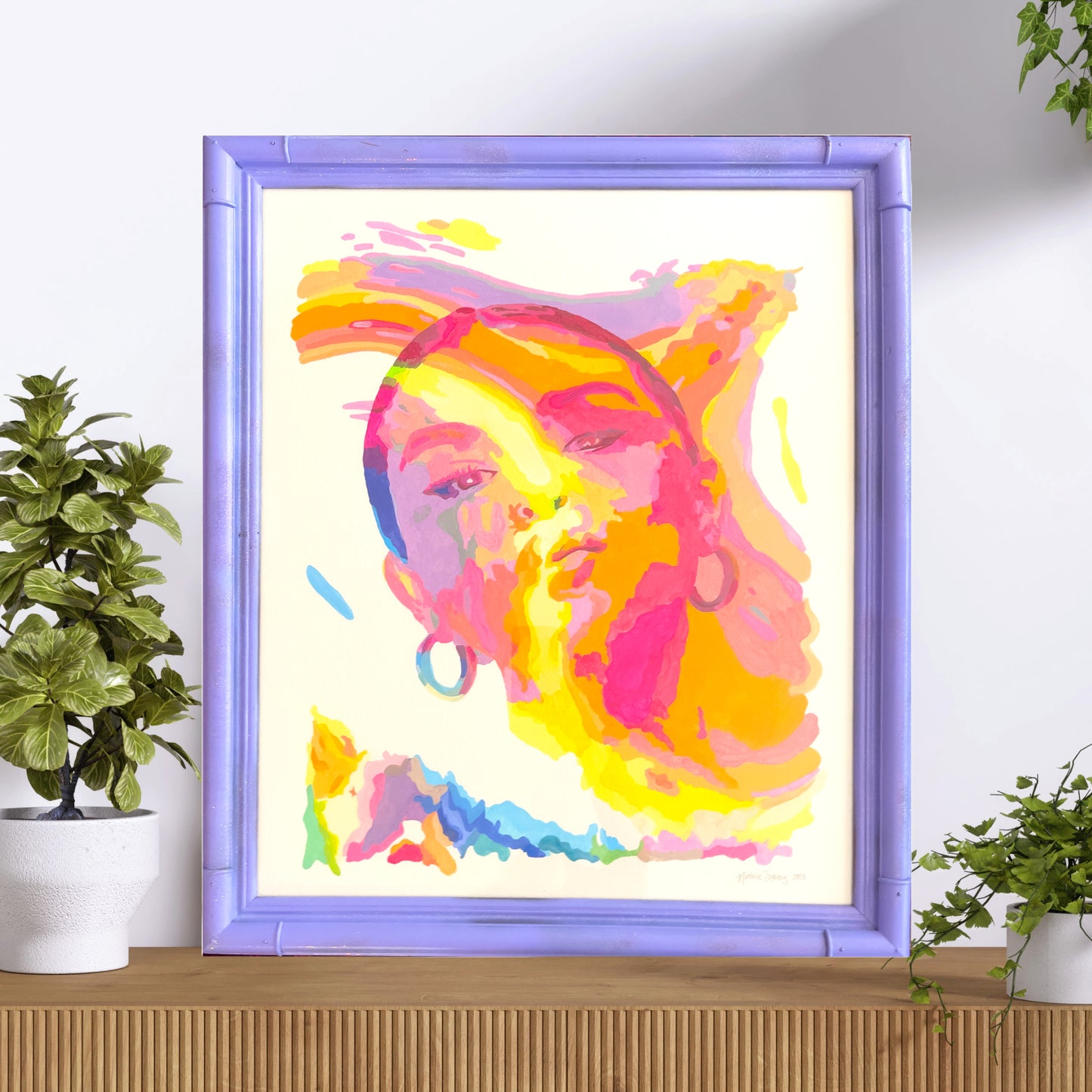Vibrant portrait painting of Selena Gomez, featuring bold and colorful brushstrokes in shades of pink, yellow, orange, and purple. The artwork is displayed in a light purple frame on a wooden surface, flanked by green plants, creating a fresh and lively presentation.