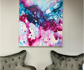 Abstract painting by Natalie Russo featuring bold splashes of pink, blue, and white, displayed on a wall between two modern gray velvet chairs. The artwork adds a vibrant and dynamic touch to the contemporary interior setting.