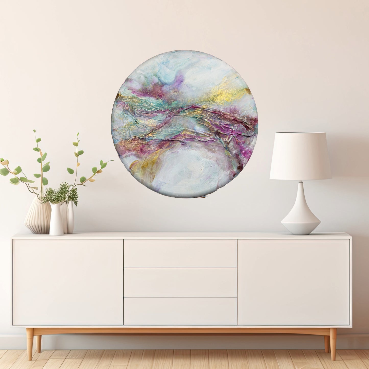 Textured round abstract painting titled 'Celestial Hues' by Natalie Russo, displayed above a modern white sideboard with a lamp and decorative vase. The artwork, featuring rich violet, gold, and blue tones with fabric texture, adds a cosmic and sophisticated touch to the contemporary interior.
